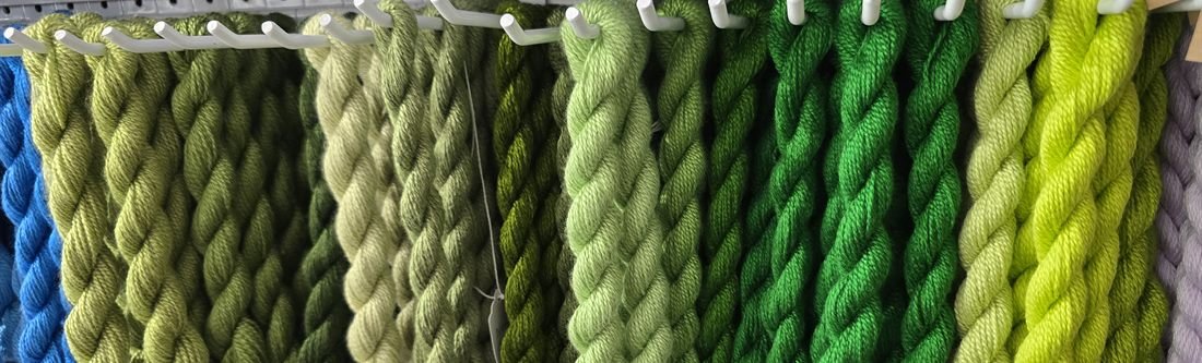 Green thread skeins hanging in rows on the wall of a needlepoint shop, running from olive through sage to bright emerald.
