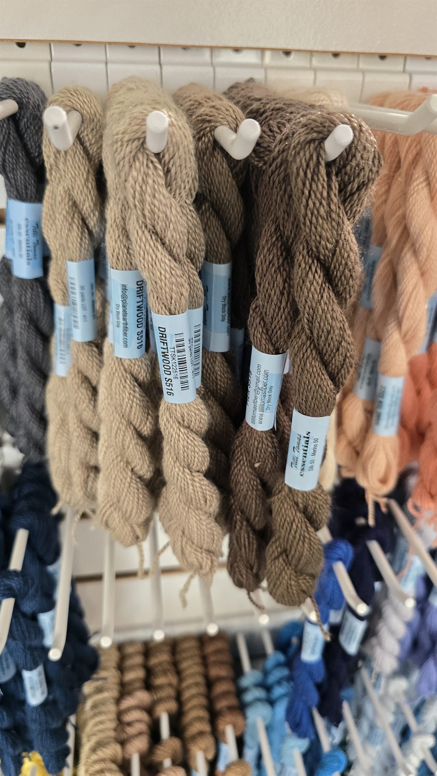 Brown and tan thread skeins hanging on a shop rack, each wrapped in a pale paper band printed with a color name, a barcode and a lot number.