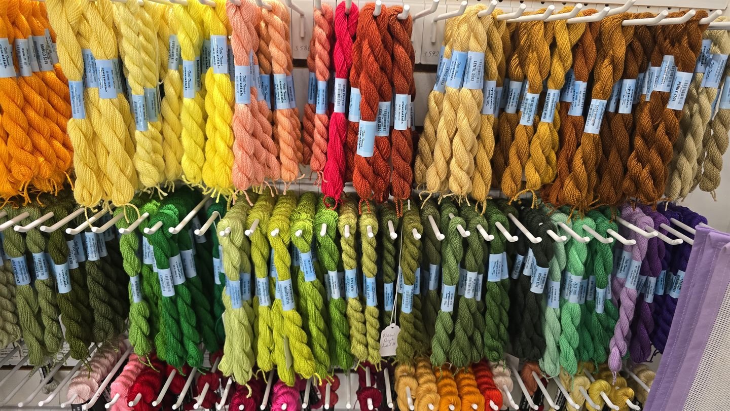 Rows of thread skeins hanging on white pegs on a shop wall, banded by color from gold and orange along the top through greens and purples below.
