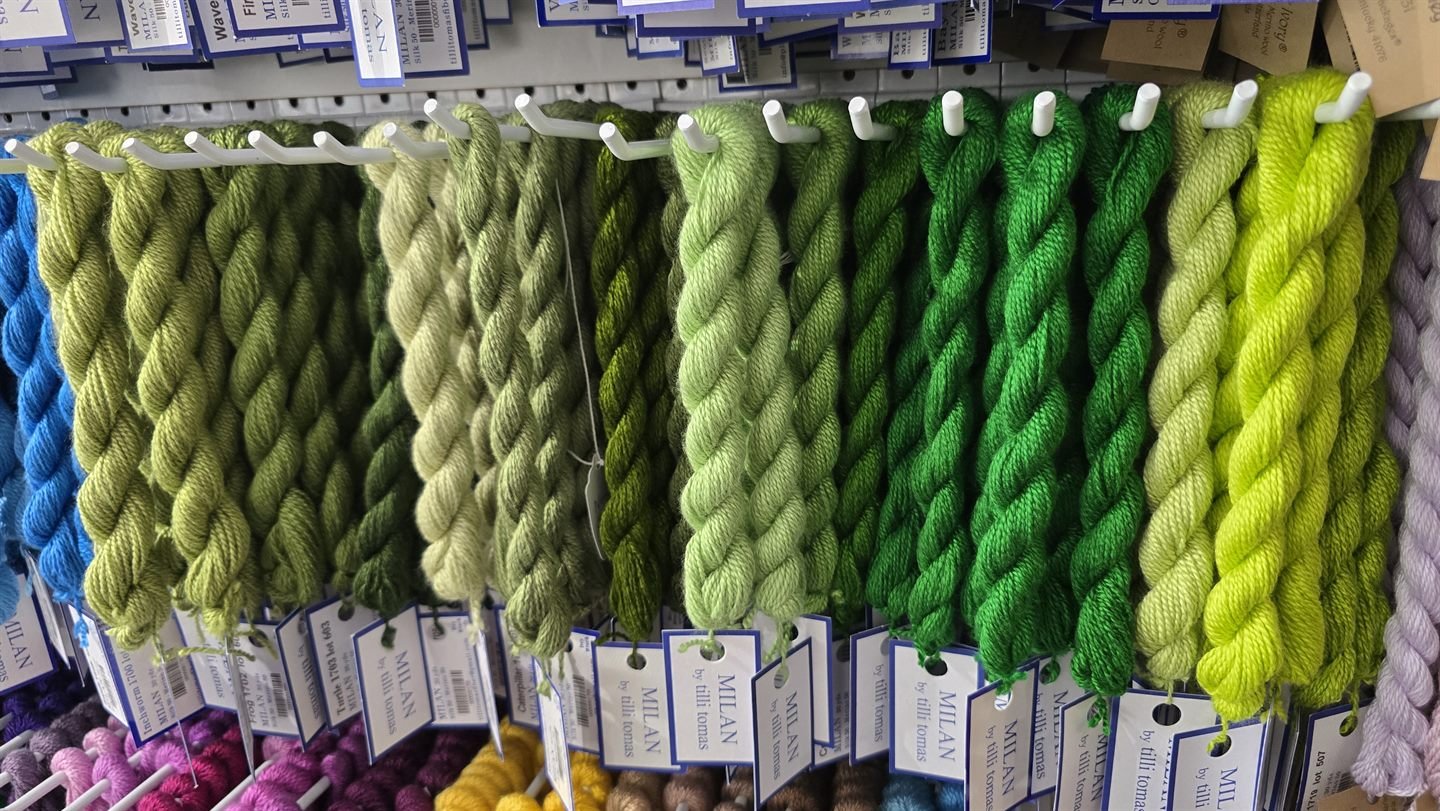 A block of green thread skeins hanging on a shop wall, running from olive and sage through deep emerald to bright chartreuse.
